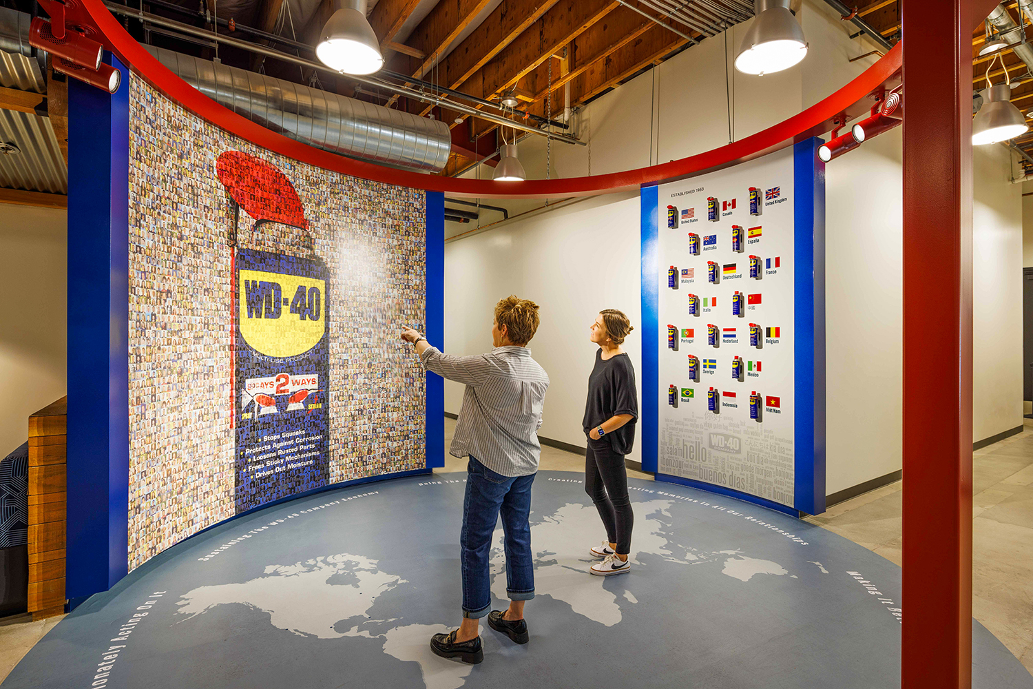 two women are looking at a graphic of a can of WD-40 made from thousands of small pictures of WD-40 employees, while in the background is a display of WD-40 cans from around the world